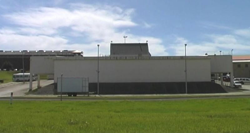 Grass field in foreground, long white block building in background with high lighting towers 